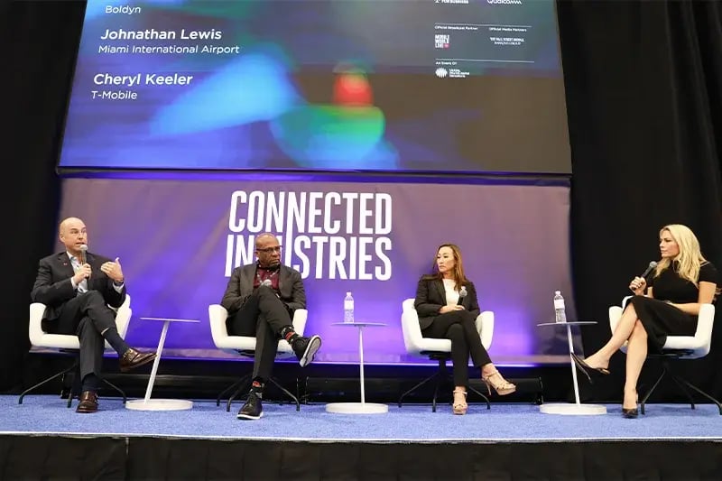 Four panelists seated on stage during a 'Connected Industries' discussion, each with a microphone, speaking in front of a large screen displaying names including Jonathan Lewis and Cheryl Keeler, with event branding in the background.