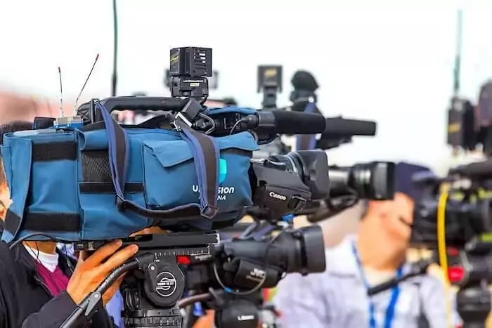 The image shows several video cameras positioned in a press zone at an event, with a focus on a prominent camera bearing the Univision logo. Multiple cameras are visible in the frame, indicating a busy media environment. The camera setup suggests a professional broadcasting or news coverage scenario, capturing footage of the event. The press area is likely dedicated to journalists covering the event.